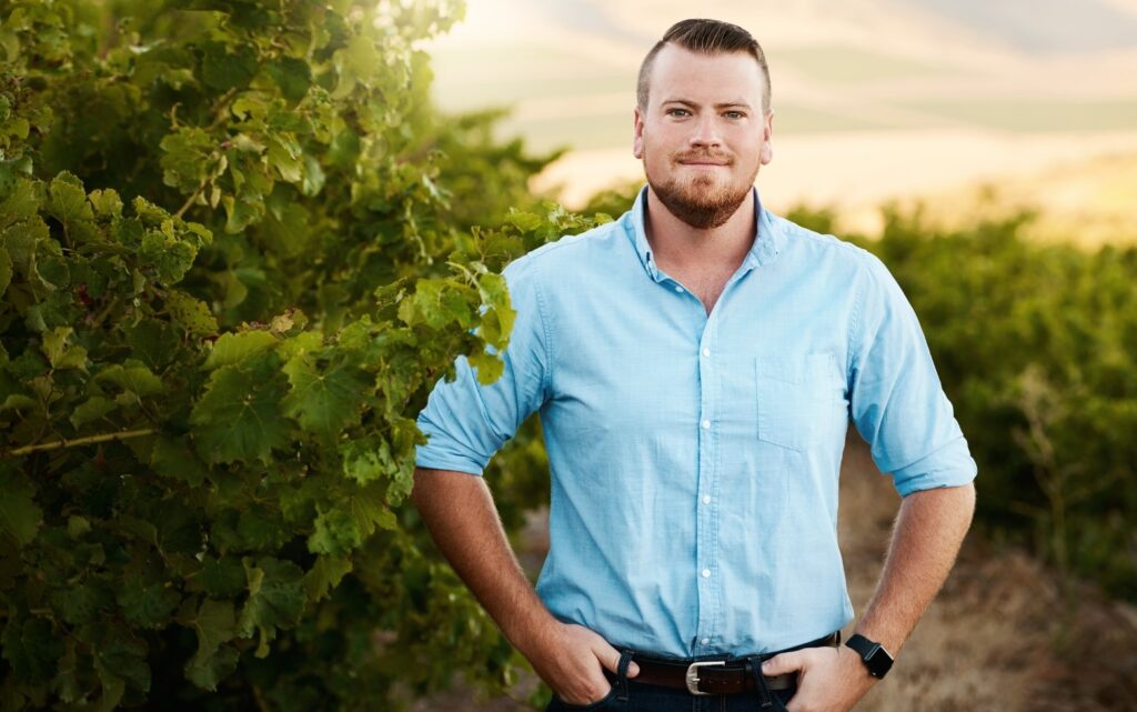 A man, wearing a blue shirt and with a beard, stands in a field amidst grapevines. He is posing for a photograph, with his hands in his pockets and a smile on his face.