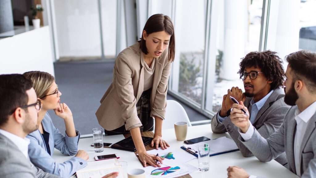 Project manager discussing plan to business workers. A woman, positioned at a table, holds a cell phone and is conversing with three seated men.