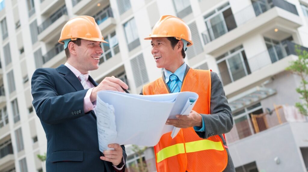 Construction workers discussing project in front of building. Two men in hard hats, one with a tie, stand in front of a building, examining a set of blueprints.